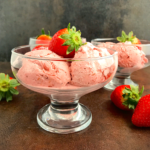 strawberry ice cream I a footed glass ice cream bowl with a strawberry on top and a second ice cream bowl at the background