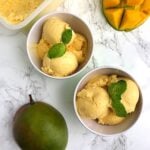 top view of mango ice cream in two green ice cream bowls surrounded by a whole mango and a cut up mango